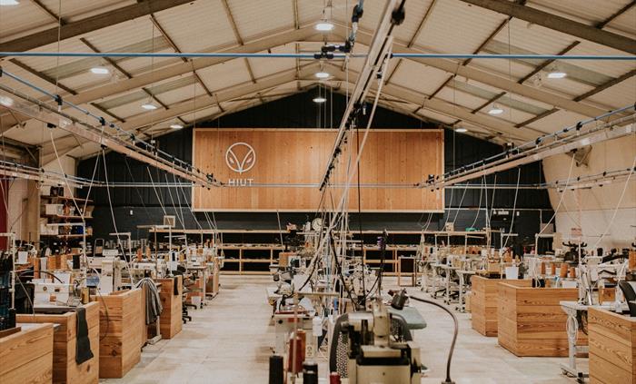 Interior of the Hiut Denim factory showing rows of wooden workstations with sewing machines under a high, well-lit ceiling. A large Hiut logo is displ